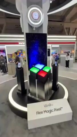 The video shows a display at a trade show featuring "LEAD 2.0 Flex Magic Pixel" technology. The display consists of a central, tower-like structure. At the top of the tower is an analog clock. Beneath the clock is a vertical screen displaying various changing patterns of color and light. At the base of the screen are nine cubes arranged in a square, each lit with a different color: blue, red, and green. The tower is mounted on a circular platform with a white light ring around the edge. Two smaller black pedestals stand on either side of the central display, each holding a smartphone-sized device displaying an image. A sign in front of the display reads "LEAD 2.0 Flex Magic Pixel." Several people are walking around in the background, looking at other displays. The environment appears to be a large, open convention hall with various booths and displays.