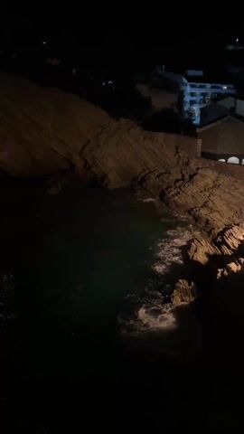 The video depicts a night scene along a rocky coast.  The sea, dark green, is being lapped by white waves. A large, layered rock formation rises from the water, partially illuminated by a light source off camera. In the distance, there are a few buildings, lit up against the dark sky.