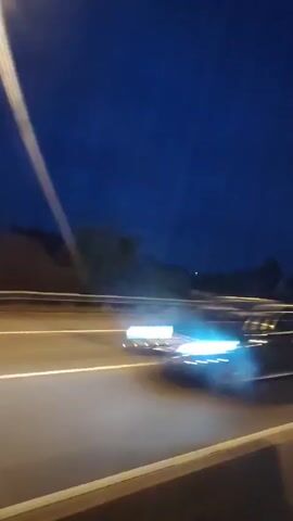 This video captures a view from a moving vehicle at night. The dark blue sky is visible above, with faint hints of distant hills and structures. A highway with illuminated lanes stretches across the foreground. Streetlights streak across the frame, creating bright lines as the vehicle passes them. Occasionally, the headlights of other vehicles create streaks of blue and white light. The overall impression is of a nighttime journey on a highway.