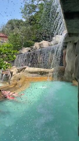 Here is a description of the video:

The video shows a constructed outdoor pool and waterfall. The pool has turquoise water. A man and a woman are in the pool, relaxing on floats. The waterfall is composed of multiple tiers with water cascading down rocks into the pool. There are lush green trees and vegetation surrounding the pool. The sky is blue, and the scene appears to be captured from under a partially covered area, as evidenced by the visible structural beams above. Water droplets are also visible on the lens of the camera.