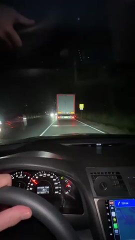 The video shows a first-person perspective from inside a car at night. The car is driving on a highway, following a large truck. The dashboard of the car is visible, showing the speedometer, tachometer, and an exterior temperature display reading 21 degrees. There is also a GPS navigation screen displayed. The road is lit by the car's headlights and the taillights of the vehicles ahead. Traffic lights and road signs are visible along the side of the road. The video is dark, and the only significant light sources are the headlights and taillights of the vehicles. The outside temperature is 21 degrees.