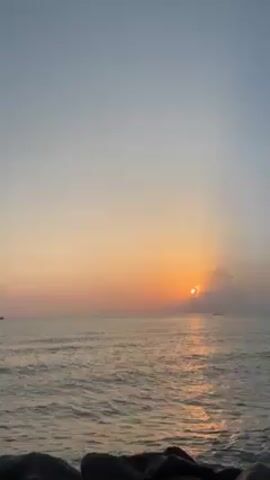 The video shows the ocean at sunrise, with the sun peeking through a cloud bank near the horizon, creating a bright reflection on the water. A few small boats are visible in the distance. In the foreground, a dark cluster of rocks or stones form the bottom edge of the frame. The sky is clear and pale blue above a gradient of orange and pink near the horizon.