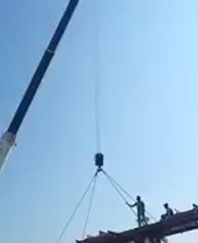 A construction crane lifts a large, heavy object while workers are on the object. The object appears to be a flatbed trailer, possibly being moved into position or assembled. The sky is clear and blue.