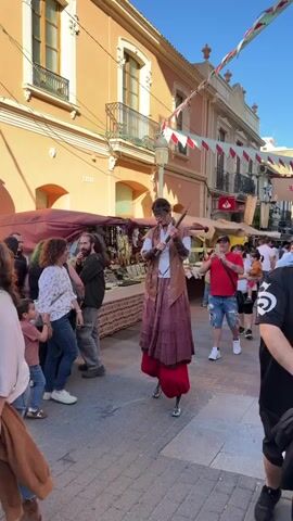 The video shows a street performer on stilts in an outdoor market. The performer, dressed in a theatrical, old-world costume with brown vest and a dark-red skirt over red pants, is also wearing dramatic makeup. They carry an axe and make deliberate movements, posing for the crowd. The crowd of people include adults and children, some taking pictures with their phones. The outdoor market is set up along a narrow street lined with light-orange buildings with balconies, and the street is adorned with pennant banners.