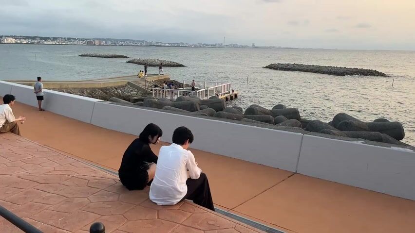 The scene shows a wide shot of a waterfront area on a cloudy day. Two people, a woman in a black dress and a man in a white shirt, are sitting on a low wall looking out at the water. A third man is sitting further to the left, also looking at the water. In the background, there are several people on a small pier-like structure. Large concrete tetrapods are piled along the shore for erosion control. In the distance, a city skyline is visible. The ground is a patterned brick surface and the wall is smooth and white. The overall setting suggests a calm, public space by the sea.