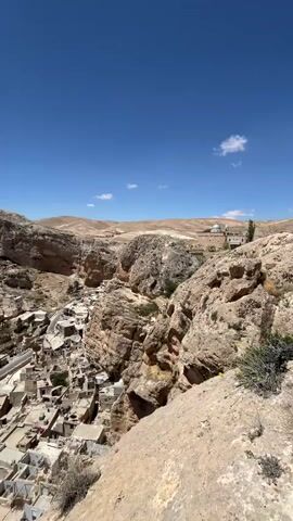The video presents a panoramic view of a town nestled within a rocky, mountainous terrain under a clear blue sky with scattered clouds. The town's architecture is characterized by densely packed buildings with flat rooftops, primarily in light, earth-toned colors. The structures are interspersed with narrow streets. The surrounding landscape consists of rugged hills and rocky cliffs, with sparse vegetation. In the background, the landscape transitions into a more open, arid expanse. A structure with a dome can be seen in the distance on the right. The camera pans slightly, offering a broader view of the town and the encompassing terrain. The foreground is dominated by rocky terrain, with the camera positioned on a high vantage point. There are sounds of people talking in the background.