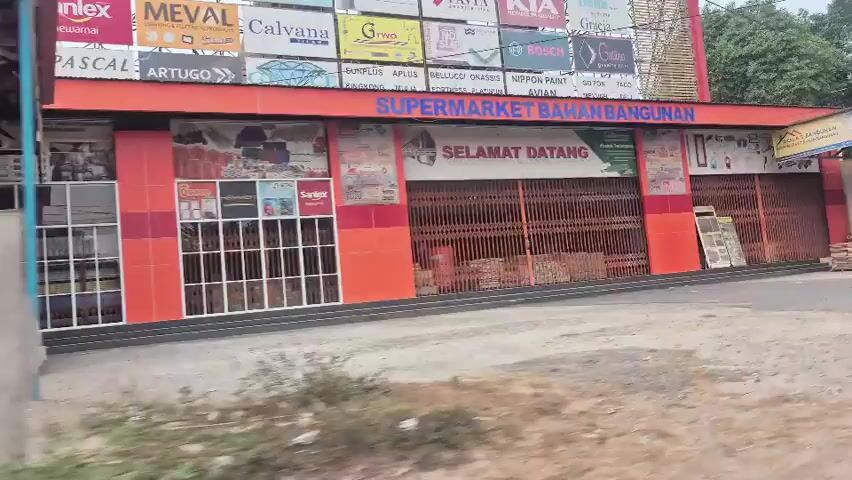 The video begins with a shot of a supermarket with the sign "SUPERMARKET BAHAN BANGUNAN" in blue letters. Red and orange shutters are closed over the entrance. Signs with various brand names such as "MEVAL", "PAVIA", "KIA", "BOSCH", "NIPPON PAINT", and "AVIAN" are visible above the supermarket. A banner inside the supermarket reads "SELAMAT DATANG" which means "Welcome". The video then pans to the right, showing the side of the supermarket and stacks of building materials. Next, the camera moves to show a rustic, wooden shack with a sign that is partially obscured. There are trees and overgrown vegetation in the background. The video continues to pan, showing more of the overgrown environment and a dirt road.