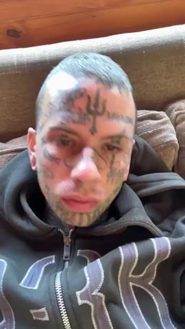A person with extensive facial tattoos and a band-aid on their nose is looking at the camera. The person is wearing a dark hooded sweatshirt with a zipper down the front. The background appears to be an indoor setting with furniture. The person makes a grimace and then opens their mouth to speak.