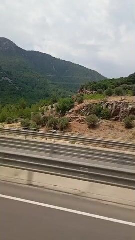 The video shows a view from a moving vehicle, looking out at a mountainous landscape. The foreground features a highway with metal guardrails. Beyond the highway, a rocky hillside is covered in green vegetation, including trees and shrubs. The sky is partly cloudy. A dark blue van is seen driving on the highway in the opposite direction of the camera.
