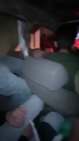 The video shows a group of people crammed into what appears to be a vehicle, possibly a car. The interior is dark. There are at least four people visible, wearing various clothing items including a green shirt, white, and black jackets. They are moving around, possibly dancing or jostling within the confined space. A flashing red light can be seen in the background through the back window. The camera view is shaky and close to the subjects.