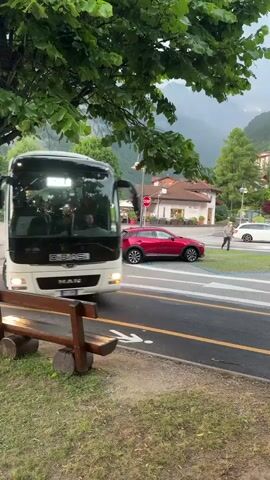 A white tour bus with blue accents, branded "DiBiASi," is driving on a road in Molveno, Italy. The bus has "DiBiASi" written in black text with a blue swoosh to the right on its side. The bus is large and appears to be a passenger vehicle for a tour group. In the background, there are mountains and trees, suggesting a natural landscape. The weather appears to be overcast, with a light mist or rain possibly falling, as evidenced by the wet appearance of the road and the leaves. The time of day is difficult to determine precisely but seems to be daytime, perhaps late afternoon or early evening, given the diffused light. A wooden bench is visible in the foreground, and a red car is driving in the opposite direction on the road. Another, smaller white bus is parked in the background. The scene evokes a sense of travel and exploration in a scenic, mountainous region.