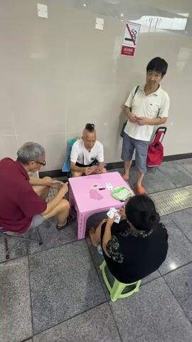 Four people are gathered around a small, pink, plastic table playing cards. One man, seated on the left, wears a maroon shirt and glasses. Opposite him, a woman in a black floral-patterned top sits on a green stool. Between them, a man with a shaved head and a mohawk, wearing a white t-shirt, is seated on a blue cushion. To the right of the table, a man in a cream-colored polo shirt and blue shorts stands, holding a red string in his hand, with a red shopping bag on wheels beside him. The scene takes place on a tiled floor with a beige-colored wall behind them. A "no smoking" sign is visible on the wall.