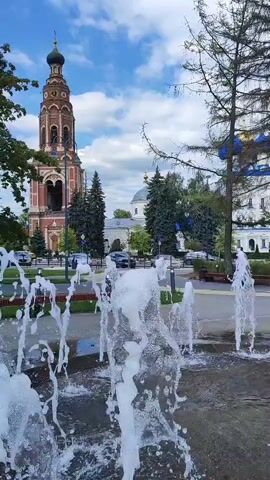 A video shows a fountain with water spraying in front of a bell tower and a church. Trees and cars are also visible. The sky is blue with clouds.