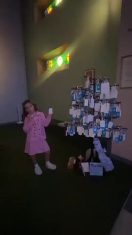 A young girl in a pink suit stands in a room with green walls and a green carpeted floor. She is holding a small white object in her right hand and her left arm is raised. To her right, there is a tree with many small blue and white tags hanging from it. In the background, there are stained glass windows and a painting on the wall. There is also a sculpture in front of the painting.