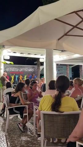 A group of people are seated at tables under a white awning. In the background, a banner reads "AZZURRA GATTEO MARE ANIMAZIONE" with a graphic of colorful figures dancing. A man in a blue shirt is speaking to the group. Several children and adults are visible, some are looking towards the man, while others are engaged in conversation or eating. The scene appears to be outdoors at night, with artificial lighting.