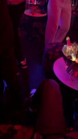 The video shows the interior of a nightclub. The lighting is dark with heavy red and blue tint. Several people are visible but they are not clearly illuminated. One person, standing near a small round table, is visible from the chest up.  This person is wearing white pants with a black belt, several necklaces and has long dark hair. They are gesturing with their hands and fingers. On the table beside them is a silver ice bucket filled with bottles. The scene is dimly lit, providing a fragmented view of the surrounding environment.