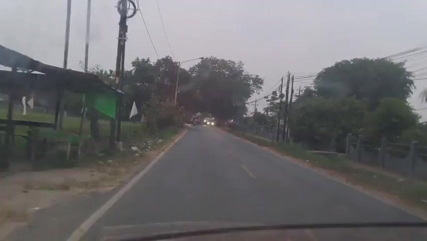 The video shows a view from inside a vehicle as it travels along a paved road. The weather appears overcast, possibly due to rain, as evidenced by droplets on the windshield and streaks from the windshield wipers. The road is flanked by vegetation, including trees and grass. There are utility poles and wires overhead, and a few houses and fences are visible on the right side of the road. Several vehicles are on the road, including cars and possibly a motorcycle. The overall scene suggests a rural or suburban setting during daylight hours.