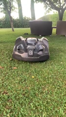 The video shows a black robotic lawnmower from the brand Husqvarna, positioned in the center of a grassy lawn. The lawnmower is dark gray with a prominent silver Husqvarna logo on its front. The surrounding environment suggests a garden or backyard setting. In the background, there are trees with lush green foliage, and two pieces of outdoor furniture, possibly a sofa and a chair, are visible. The overall scene appears to be outdoors, with natural light indicating it's daytime. The weather seems pleasant and sunny. There are no people visible in the video. The activity captured is the static presence of the robotic lawnmower, possibly indicating it is paused or awaiting activation. The camera is positioned low, at the level of the lawnmower, and slightly angled upwards. The footage is recorded in a slightly shaky manner, suggesting it might be handheld. There is no discernible text other than the brand logo on the lawnmower. The location context provided mentions Castiglione d'Orcia, Italy, which hints at a possible European landscape with its characteristic cypress trees often found in the Tuscan region.