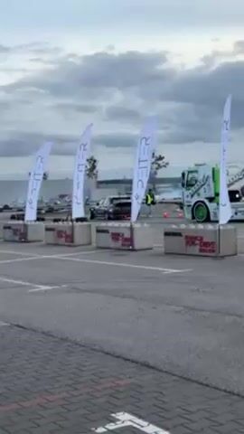 The video shows a car drifting on an asphalt surface, against a backdrop of a cloudy sky.  Four white flags, each bearing the vertical word "PEITER," stand in a row, anchored in concrete blocks that are also marked with advertising. A green and white truck is parked near the flags on the right. A person in a yellow vest stands near orange cones. The car leaves a trail of smoke as it drifts. Other cars are parked further in the background.