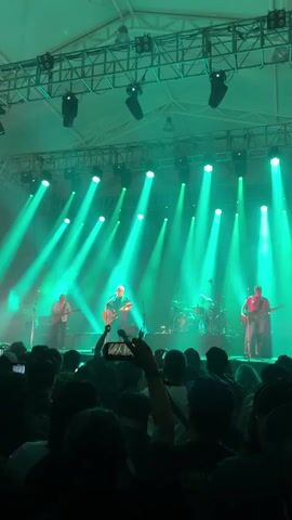 A live performance is underway on a stage lit with numerous green spotlights. Four musicians, including a drummer, are visible performing in front of a crowd. The audience is densely packed, with many people holding up phones to record the show. The venue appears to be an indoor tent structure, supported by metal beams.