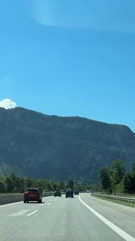 A sunny day with a clear blue sky is depicted, with a mountain range in the background. A highway with several cars driving on it is in the foreground. The cars are moving away from the camera. The road is paved and has lane markings. Trees and foliage are visible on the sides of the road.