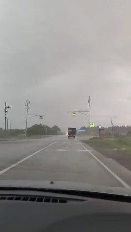 The video shows a driver's perspective while driving on a wet road in heavy rain. A truck is driving ahead in the same direction. The wipers are on, working to clear the windshield. Pedestrian crossing signs are visible on either side of the road. The sky is overcast and gray.