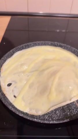 The video shows a close-up shot of a crêpe being cooked in a frying pan. The crêpe is a light yellow color with some golden brown spots beginning to form. A metal utensil with a white spatula attachment is used to lift and fold a portion of the crêpe. The background shows a black stovetop and a tiled wall. The lighting is warm and focused on the cooking crêpe.