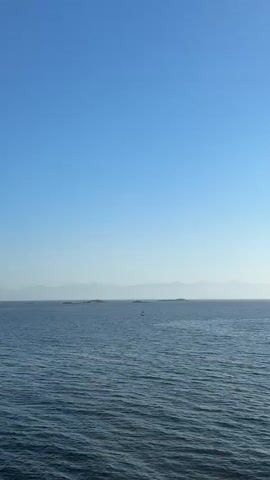 The video shows a view of the ocean with some islands and mountains in the distance. The sky is a clear blue, and the water is a deep blue with gentle waves. As the video progresses, the camera pans to the right, revealing more islands with trees and houses on them.