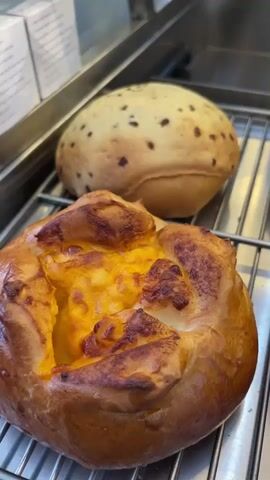 This is a close-up, vertical, eye-level shot of a bakery display. The focus is on a cooling rack filled with various baked goods. In the foreground, to the left, is a golden-brown pastry with a cut-out center filled with what appears to be melted cheese or a creamy orange filling. To its right is another similar pastry, slightly out of focus. Behind these, on the upper rack, are two round buns. The one on the left is pale with dark speckles scattered across its surface, resembling chocolate chips or seeds. The bun on the right is also pale and has a slightly textured, golden-brown, crispy topping. Another similar bun with a textured topping is visible on the right side of the upper rack, partially obscured. The background is a metallic surface, likely part of the display case. The lighting is bright and even, highlighting the textures and colors of the baked goods.