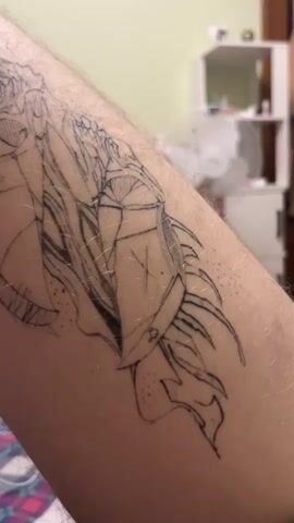 A close-up video shows an unfinished tattoo on a person's arm. The tattoo appears to be of a dragon or mythical creature with intricate details, including scales, wings, and sharp claws. The lines are still visible and some shading has been applied, but the tattoo is clearly not complete. The arm is hairy and the skin is pale. In the background, a room is visible with shelves and a fan. The lighting is soft and natural.