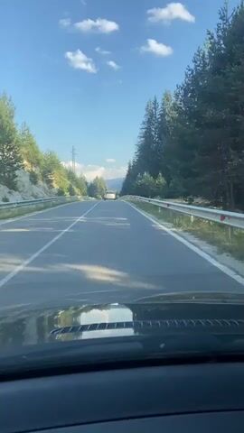 The video shows a first-person perspective of a car driving on a paved road. The road is lined with guardrails on both sides and is surrounded by a forest of tall evergreen trees on the right and a grassy slope with some trees on the left. The sky is blue with scattered white clouds. There is another white car in the distance ahead. The dashboard of the car is visible in the foreground.