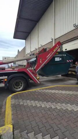 The video captures a daytime scene in an industrial or commercial area in Leudelange, Luxembourg. A large red truck, equipped with a hook-lift system, is in the process of lowering or adjusting a substantial green skip/dumpster. The truck has a grey cab and black tires.

A man, dressed in dark work attire and a high-visibility vest, stands near the back of the green skip, observing the operation. The skip is positioned next to a large building with white corrugated metal siding and a grey overhang, indicative of a warehouse or factory. The ground is paved with interlocking bricks, featuring yellow painted lines. The sky is overcast.

Visible text includes "B.P.A.T" and "BOMAREL" on the red hook-lift arm. The green skip also has "B.P.A.T." with a logo, the number "8311", and a phone number "TEL : 26 59 71 19". The activity appears to be a routine waste management or container handling process.