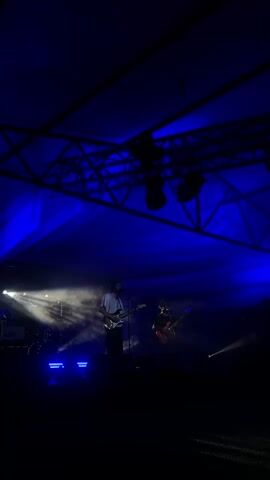 A dimly lit outdoor concert stage is illuminated by blue spotlights. Two men are visible. The man on the left is standing center stage, playing a guitar and singing into a microphone. He is wearing a light-colored shirt and dark pants. The man on the right is further back on stage, also playing a guitar, wearing dark clothing. Smoke or haze drifts across the stage, catching the light. The ceiling of the stage area appears to be a large, taut fabric structure, with exposed metal trusses and lighting equipment overhead. The overall atmosphere is dark, with blue light dominating the scene.