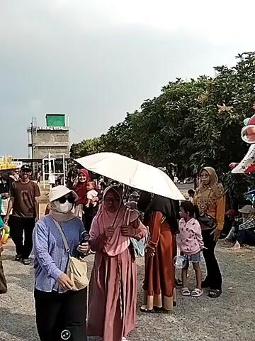 The video shows an outdoor scene with many people gathered. There are several stalls selling balloons and toys. One stall has a large display of colorful balloons shaped like animals and an airplane. People are seen walking around, some are holding umbrellas, and others are wearing masks. The environment appears to be a park or an open area with trees and some buildings in the background. The overall atmosphere seems to be festive, likely a local event or gathering.