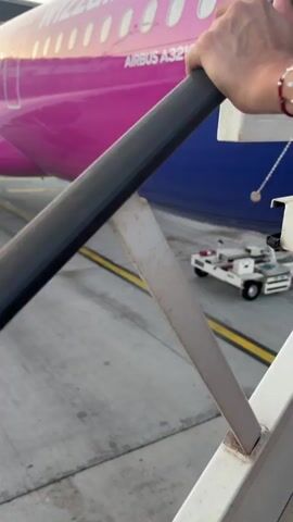 The video shows a person boarding an airplane via stairs. The airplane is pink and blue with the text "Wizz Air" and "Airbus A321neo" visible on its side. A baggage tug is parked behind the stairs, and the ground is paved. The camera pans down the stairs, showing the metal steps and railings. The person boarding is wearing white shoes. The sun is shining, casting shadows on the ground.
