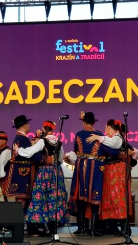 This video captures a folk dance performance on a stage in Prešov, Slovakia. A group of dancers, dressed in traditional Slovak costumes, are engaged in a lively performance. The costumes are vibrantly colored and richly decorated. The women wear floral print skirts and white blouses, topped with embroidered vests and red accents. The men are adorned in dark vests, white shirts, and black hats, with similar elaborate embroidery.

The stage backdrop is a large purple screen displaying the text "1. ROČNÍK FESTIVAL KRAJÍN A TRADÍCII" in colorful, stylized lettering, followed by the large yellow word "GADECZANIE". This indicates the event is the first annual festival celebrating lands and traditions. The dancers are positioned in a line, some in pairs, in what appears to be a choreographed dance. Microphones on stands are visible in front of some of the performers, suggesting a live musical accompaniment or vocal performance.

The lighting suggests an indoor event or evening performance, with bright stage lights illuminating the scene. The dancers' expressions convey joy and energy, hinting at the celebratory nature of the festival. The overall atmosphere is festive and cultural, highlighting Slovak traditions. The presence of stage equipment, such as lighting rigs and speakers, further confirms the setting as a performance venue.