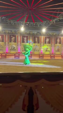 A woman in a green outfit is dancing in a dirt arena. She is wearing a green skirt and a green top, and she has a green scarf that she is waving around. The arena is decorated with murals of ancient Greek or Roman scenes, and there are lights strung across the ceiling. She is performing a solo dance.