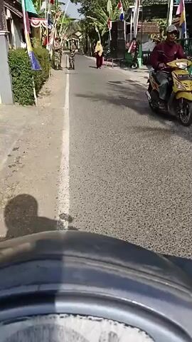 The video is captured from the perspective of someone riding a motorcycle. The road ahead is paved and appears to be a village street, lined with houses and vegetation. Several individuals are visible. On the left side of the road, two people in camouflage uniforms are standing, holding what appear to be flags or poles. Further down the road, a person in a yellow hijab and a dark red dress is walking away from the camera. To the right of this person, another individual in a camouflage uniform is walking. The atmosphere seems festive, with Indonesian flags displayed along the street. The lighting suggests it is daytime, with sunlight casting shadows on the road. The motorcycle's dashboard is partially visible in the foreground.