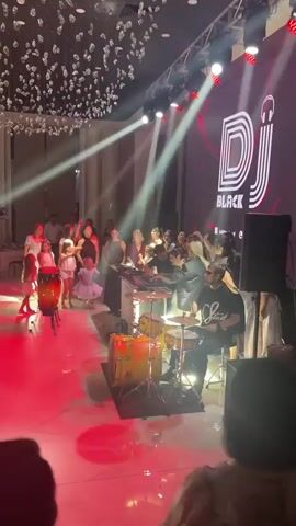 The video shows a lively indoor event, likely a party or reception, with a DJ set up on a raised platform. A large screen behind the DJ displays the logo "DJ BLACK" with a headphone icon. Various lighting effects, including spotlights and colored beams, illuminate the scene. In the foreground, to the left, are two conga drums. To the right of the DJ booth, a drummer is playing a drum kit. A crowd of people, including adults and children, are gathered around the DJ and dancing. Several individuals are holding up their phones, presumably recording or taking pictures. The floor appears to be a polished marble or tile. The ceiling is decorated with numerous small, hanging white elements that resemble small flowers or crystals, catching the light. The overall atmosphere is energetic and festive.