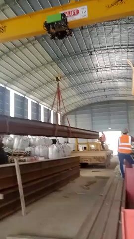 A yellow overhead crane with the words SAFETY FIRST and a logo is moving a long, dark brown metal beam. The beam is suspended by red straps attached to the crane's hook. The crane is inside a large, industrial building with a corrugated metal roof and natural light coming from windows along the sides. Below the beam, stacks of similar metal beams and large white sacks are visible. To the right, a flatbed truck is parked. In the background, a person wearing an orange vest is standing near the truck. The crane is moving from left to right.
