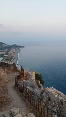 The video shows a slow pan across a rocky cliff overlooking a calm, blue ocean. The sky is clear and transitioning from a soft pink and orange at the horizon to a pale blue overhead, suggesting either sunrise or sunset. A metal railing runs along the edge of the cliff path, providing a safety barrier. The path itself is made of dirt and scattered rocks. In the foreground, on the cliff, are remnants of stone structures, possibly ancient ruins, partially overgrown with dry grass and small bushes. The water is smooth with very gentle ripples, and the coastline is visible in the distance to the left, appearing hazy. The camera movement is smooth and steady, creating a peaceful and scenic atmosphere. There are no people or animals visible in the video.