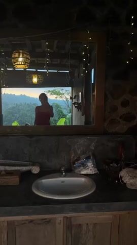 A person in a red swimsuit takes a mirror selfie in what appears to be an outdoor bathroom or changing area. The setting looks tropical with lush green foliage visible in the background through an open doorway or large window. The time of day appears to be sunset or sunrise, indicated by the warm glow in the sky. The bathroom itself has a rustic aesthetic with natural materials like wood and stone. There is a sink in the foreground, and decorative lights hang from the ceiling. The person is posing with one hand on their hip.