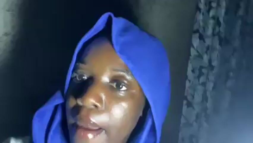 A person with dark skin, wearing a blue hooded sweatshirt, is looking directly at the camera. They appear to be indoors, possibly in a dimly lit room, with a wall visible in the background. The person briefly holds up a white container with a green top. The camera then rotates, showing the person lying down, still looking at the camera and smiling. A black and white patterned fabric is visible in the background.