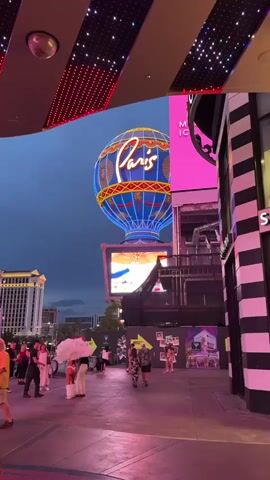 The video captures an outdoor urban scene at dusk. A large, illuminated hot air balloon with the word "Paris" emblazoned on it is prominently displayed against a cloudy sky. Below the balloon, a large screen displays an image. The foreground features a walkway with people strolling and interacting. To the right, a building with black and white striped facade is visible. In the background, large hotel-like buildings with illuminated windows are present, suggesting a commercial or entertainment district. The ground reflects the ambient pink and red lighting. The overall atmosphere appears lively, with a mix of architectural structures, signage, and pedestrian activity.