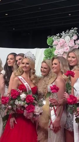 A group of women is posing for a photo. One woman, wearing a red dress, a crown, and a sash that says "MISS DEN," stands in the center holding a bouquet of pink and red flowers. Other women surround her, some holding single red roses. Most of the women have blonde hair and are wearing formal dresses. A large floral arrangement of white and pink roses sits to the right. The background is a room with a dark ceiling.