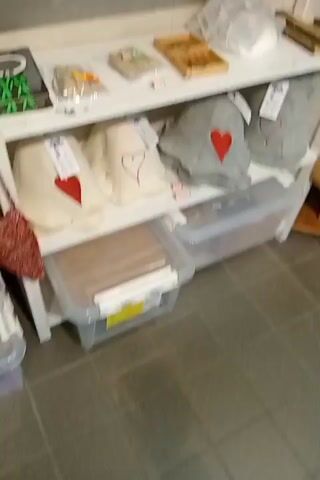 The video shows shelves stocked with various items, possibly in a gift shop or souvenir store. On the top shelf, there are boxes of products, some with yellow and brown packaging. The second shelf displays more boxed items and some items in clear plastic bags, along with a wooden tray containing small objects. The third shelf holds white fabric items decorated with red hearts, a grey garment with a red heart, and clear plastic storage bins underneath. The fourth shelf has more fabric items and storage bins containing brown items. The fifth shelf is filled with plush toys, including stuffed animals and a stuffed seal. Next to the plush toys are rows of small dark bottles. The sixth shelf is laden with numerous colorful boxes of what appear to be chocolates or candies. The seventh shelf holds more boxed items. The eighth shelf has plush toys, including cats, dogs, and a duck. The ninth shelf contains white shelves with more stuffed animals and what appear to be decorative wooden carvings. Further to the right, on another set of shelves, are mugs with various designs and small decorative items. In the foreground, a rack displays brochures or pamphlets with the word "Finland" visible on some of them. There are also hats with "FINLAND" printed on them, along with other hats and a brown hat on a higher shelf. Several plush dogs and a stuffed husky are also visible. The overall impression is of a retail environment selling a variety of goods, with a noticeable theme of Finland merchandise.
