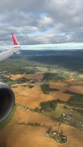 The video shows an aerial view from an airplane window. The wing of the plane, with a red logo, is visible in the top left corner. Below, a rural landscape unfolds with fields of golden crops, patches of green forest, and scattered houses and buildings. The sky is partly cloudy, with sunlight casting shadows on the ground. The video appears to be shot from a moving aircraft.