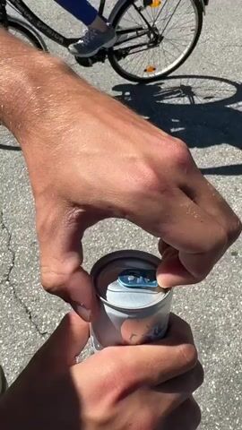 The video shows a close-up of a person's hands opening a can. The left hand holds the can, while the right hand uses a thumb to lift the tab on the can's lid. The tab is blue. The can is silver and appears to be a beverage can. The background is a grey, textured surface, possibly asphalt, suggesting an outdoor setting. The lighting is bright, indicating daylight. The action is focused on the process of opening the can.