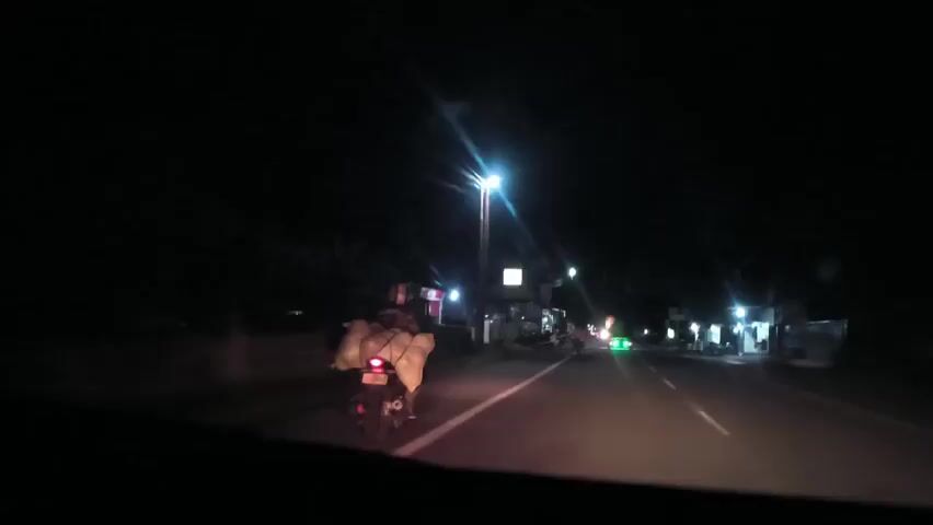 A vehicle is driving down a paved road at night. The road is illuminated by streetlights. There are buildings on either side of the road, many appearing to be small shops. Ahead, a motorcycle carries a large load secured to the back. The motorcycle's taillight is visible. Other vehicles are seen in the distance, moving along the same road.  The scene is dark, with the headlights and streetlights providing the main sources of illumination.
