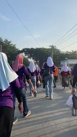 A group of people, predominantly women wearing hijabs and purple shirts, are walking in a procession along a paved path. The sky is clear and the lighting suggests it is daytime. The people appear to be moving away from the camera. Some individuals are carrying bags. The environment includes trees and utility poles. The perspective is from behind the group, capturing their movement and the surrounding area.